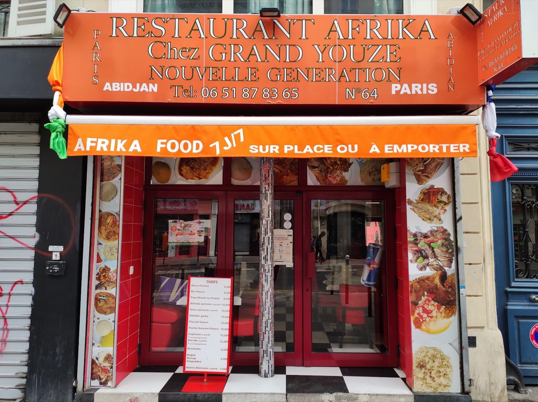 La devanture de Chez Grand Youze au 64: enseigne orange, Abidjan et Paris écrits aux deux bouts, store orange, drapeaux noués